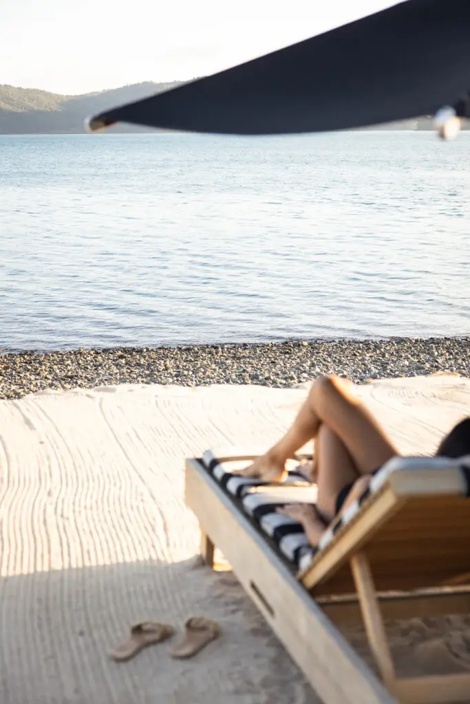 A white-sand beach by still waters with islands in the backdrop. A white woman's leg can be seen on a striped beach lounger.