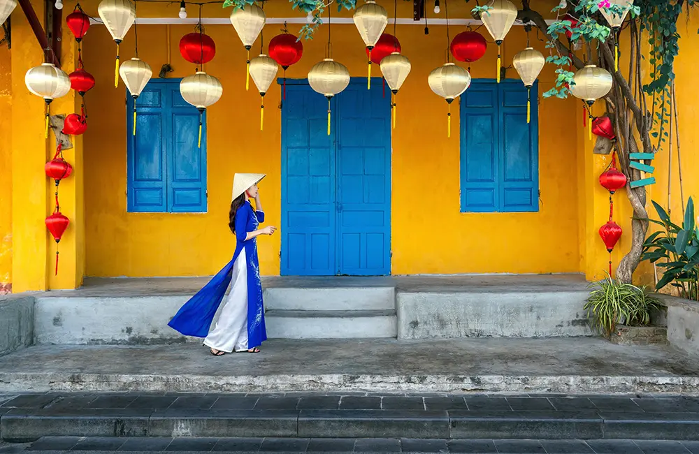 Woman in traditional Vietnamese dress walking past bright building in Hoi An, Vietnam.