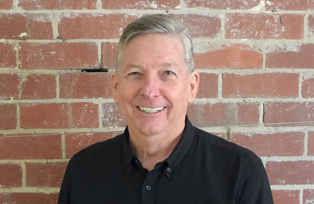 Movers + Shakers: Katrina Barry joins ATIA board; Shelley Beasley steps down 2 Corporate headshot of Tom Manwaring in black shirt against brick backdrop.