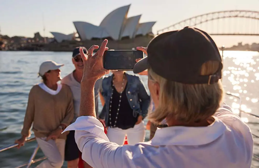 Woman taking a photo of passengers on a cruise ship in Sydney Harbour with the Sydney Harbour Bridge and Opera House in background.