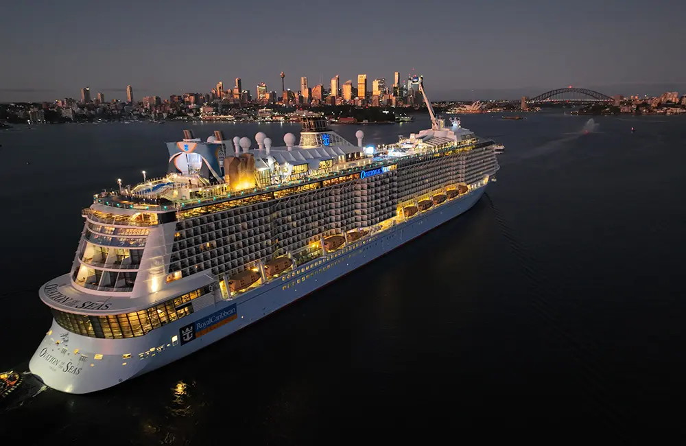 Standing ovation: Royal Caribbean ship sails into Sydney for record summer season 2 Ovation of the Seas enters Sydney Harbour at night with city skyline and bridge in background.