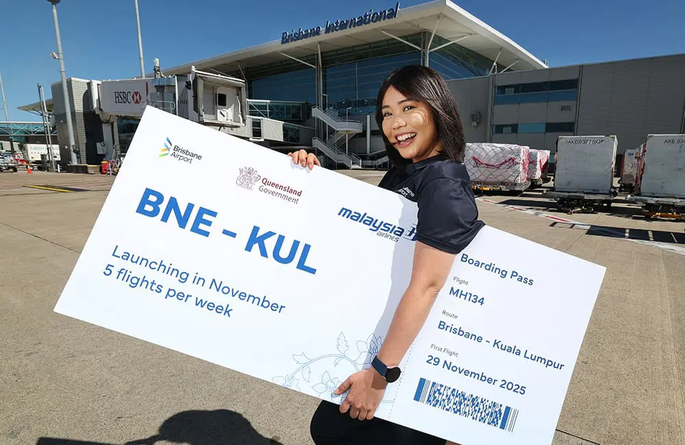 BNE back to 33 direct international destinations with return of Asian airline & route 1 Female Brisbane Airport staff member holds oversized Malaysia Airlines BNE-KUL ticket on tarmac.