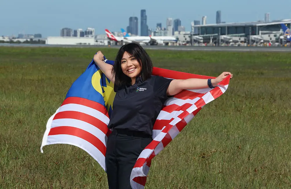 BNE back to 33 direct international destinations with return of Asian airline & route 3 Female Brisbane Airport staff member holds Malaysia flag in field with International Terminal in background.