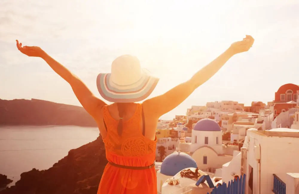 When life gives you $100K to spend, 13% of Aussies say "Book the trip!" 1 Woman with back to camera and arms outstretched in Santorini, Greece at sunset. $100K to spend