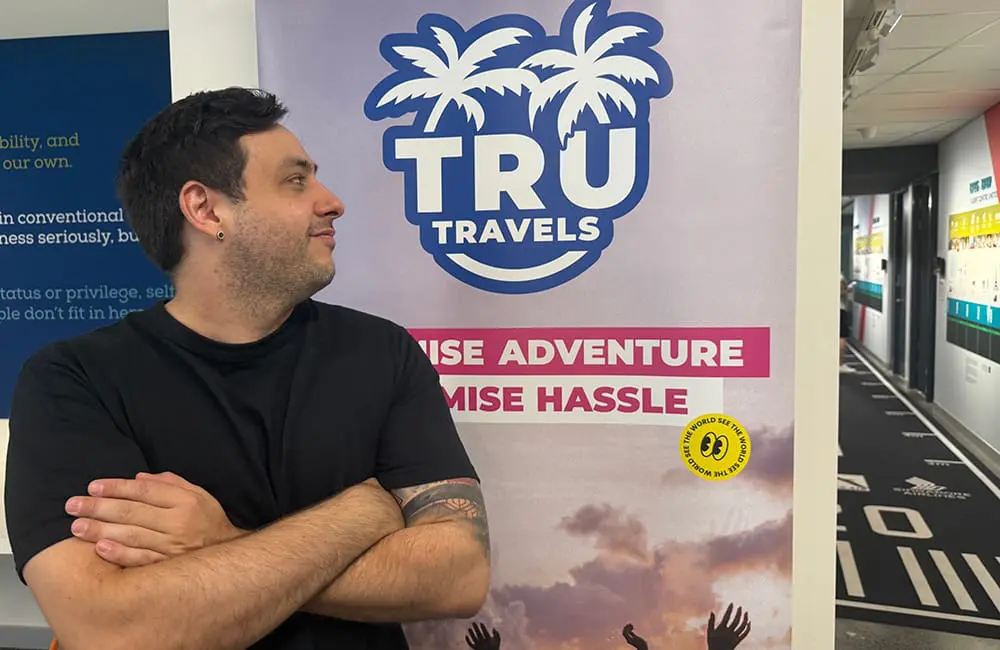 Movers + Shakers: TruTravels establishes first-ever Australia-based reservations department helmed by res guru Drew Appleton 1 Man in black t-shirt looks at TruTravels banner within a travel agency store.