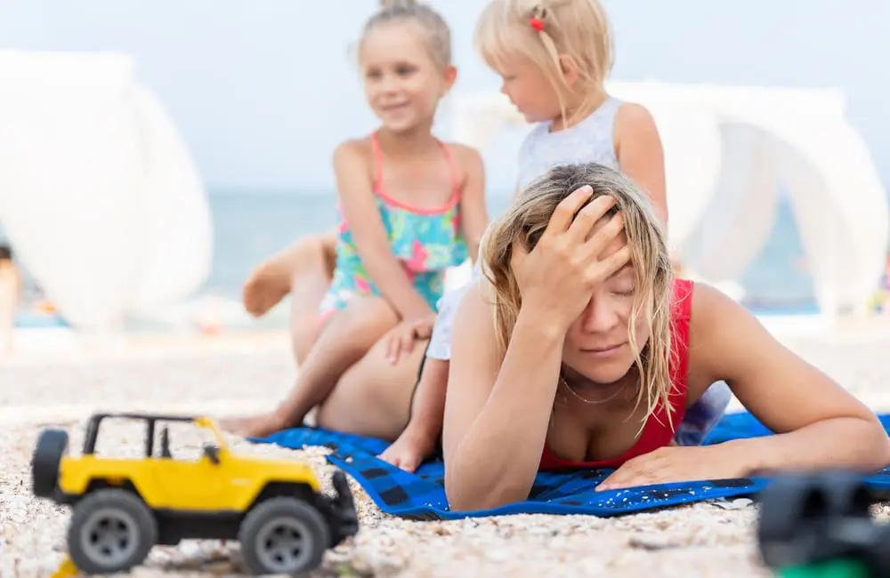 Stressed mum lying on beach with toddlers climbing on her back during overseas trip. Image: Shutterstock