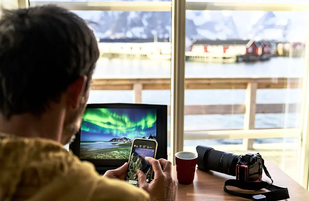 Expedia TAAP rolls out smarter payment options to boost agent revenue & efficiency 1 Man travel advisors looking at map on smartphone with DSLR camera and Northern Lights image on screen in Norway.