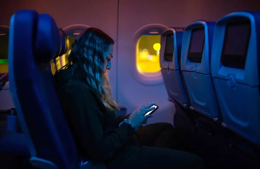 Most Aussies say this is the best time to fly overseas – and it’s not what you’d expect 1 Female plane passenger on smartphone during late-night flight – preferred times for flying overseas. Image: Shutterstock