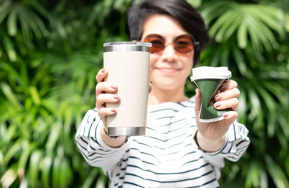 Holiday mode activated? Why we forget to pack our green habits and how travel can close the gap 2 Female traveller holding disposable coffee cup and reusable drink vessel – holiday mode sustainability