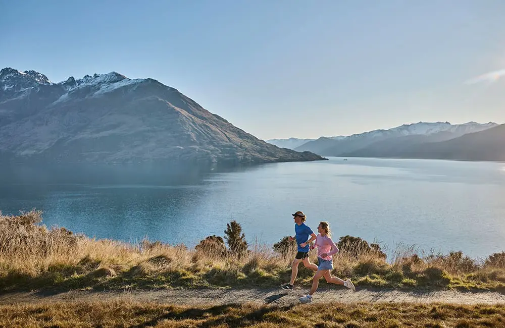 Couple running outdoors past a lake in Queenstown, Otago – Tourism New Zealand Active Escapes campaign.  Image: Camilla Rutherford