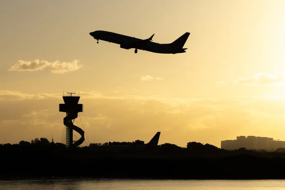 A Virgin Australia Boeing B737 MAX 8 plane, registration VH-8IA, taking off from Sydney Kingsford Smith Airport as flight VA404 to Adelaide. In the left background is the air traffic control tower. In the centre is a Qantas Boeing B737-838 plane, registration VH-VZU, taxiing to the international terminal after arriving as flight QF162 from Wellington. This image was taken near Kyeemagh Beach, Botany Bay on a sunny morning shortly after sunrise on 14 March 2026.