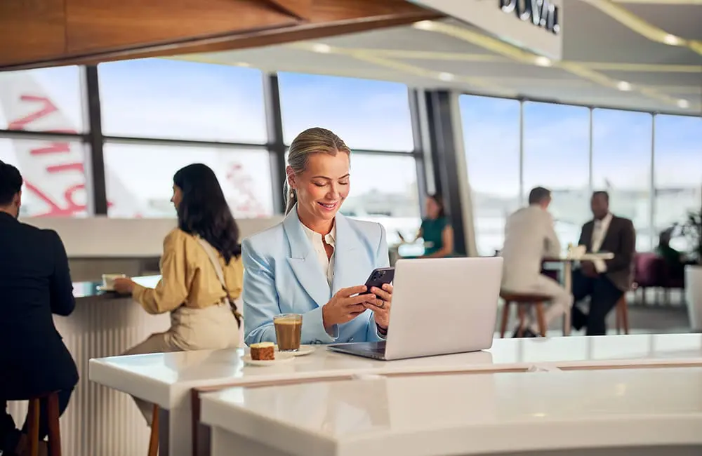 Virgin Australia is only Aussie airline to offer free same-day flight changes for Economy Flex flyers 3 Businesswoman in Virgin Australia lounge with smartphone and laptop with aircraft in background
