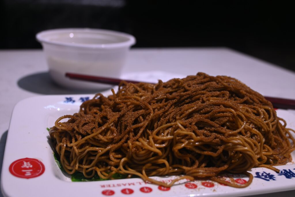 A white plate with noodles covered in brown sauce and sprinkled heavily in dried shrimp roe