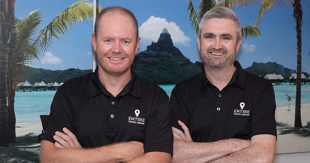 Two men wearing branded Entire Travel Group black shirts stand in front of South Pacific image backdrop.
