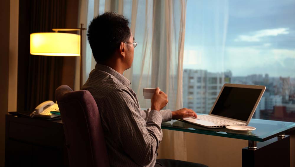 Businessman in hotel room looking out of window from desk for corporate travel hacks concept
