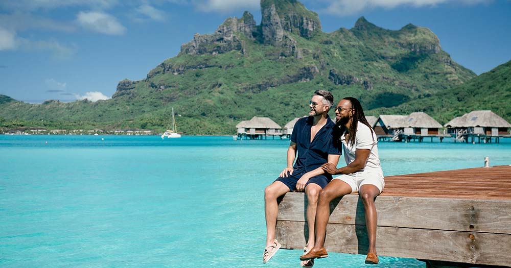 The lowdown from Tahiti as destination unveils biggest-ever incentive 1 Male couple sit on boardwalk overlooking ocean in Bora Bora in the Island of Tahiti with overwater bungalows and mountains behind.