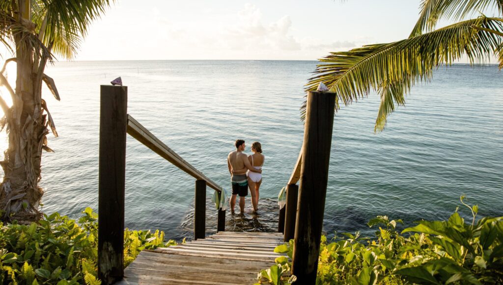 A couple standing arm in arm at the end of a jetty, leading to a calm blue waters