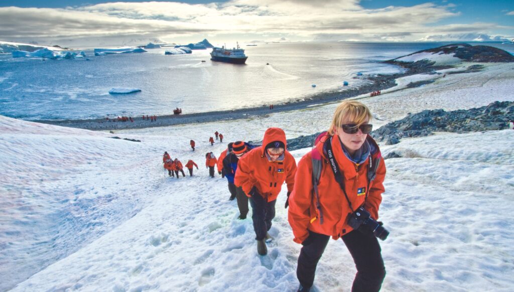 A row of red-coat wearing travellers walking up a mountain-covered slope with a bay and cruise ship in the distance.