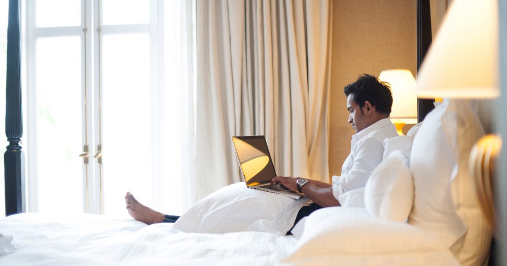 ATO targets bleisure trips: Corporate Traveller warns Aussies to tread carefully this tax season 2 Young businessman in hotel room using laptop in bed for bleisure travel trip.