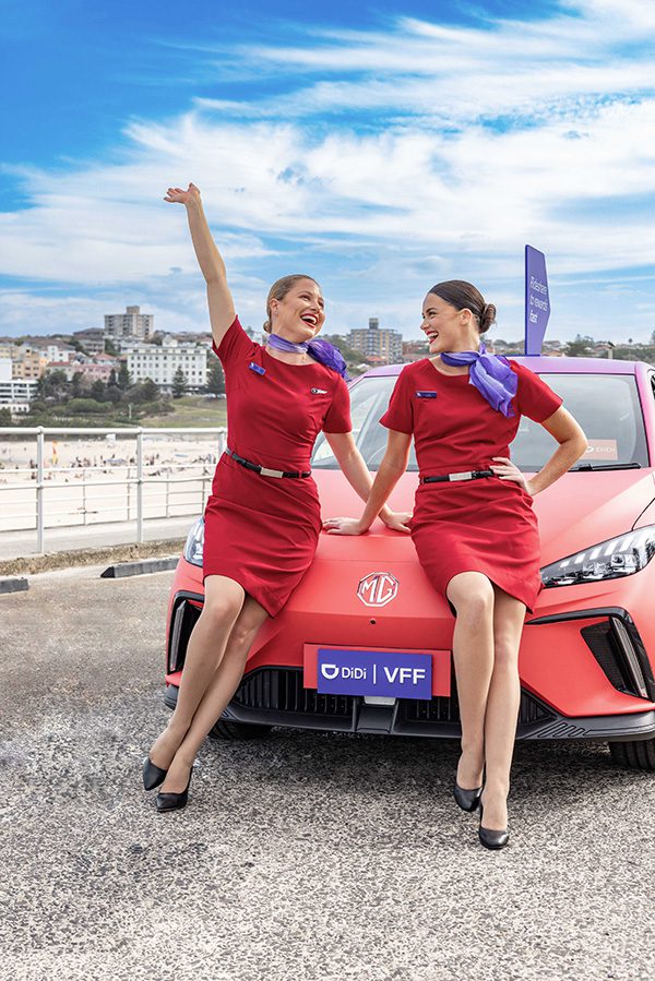 Two Virgin Australia flight attendants sitting on bonnet of branded red DiDi car at Bondi Beach.