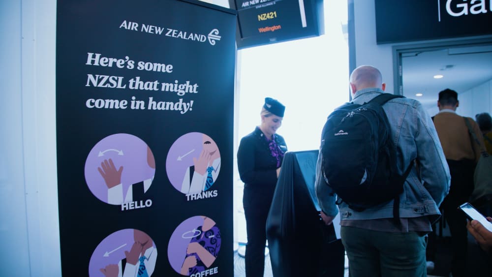 Sign reading 'Here's some NZSL that might come in handy!' displaying sign language to say hello, thanks and coffee. One flight attendant and one traveller stand at the Air NewZealand boarding gate.