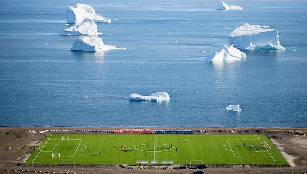 Green football pitch on Disko Island, Greenland with icebergs floating in sea.