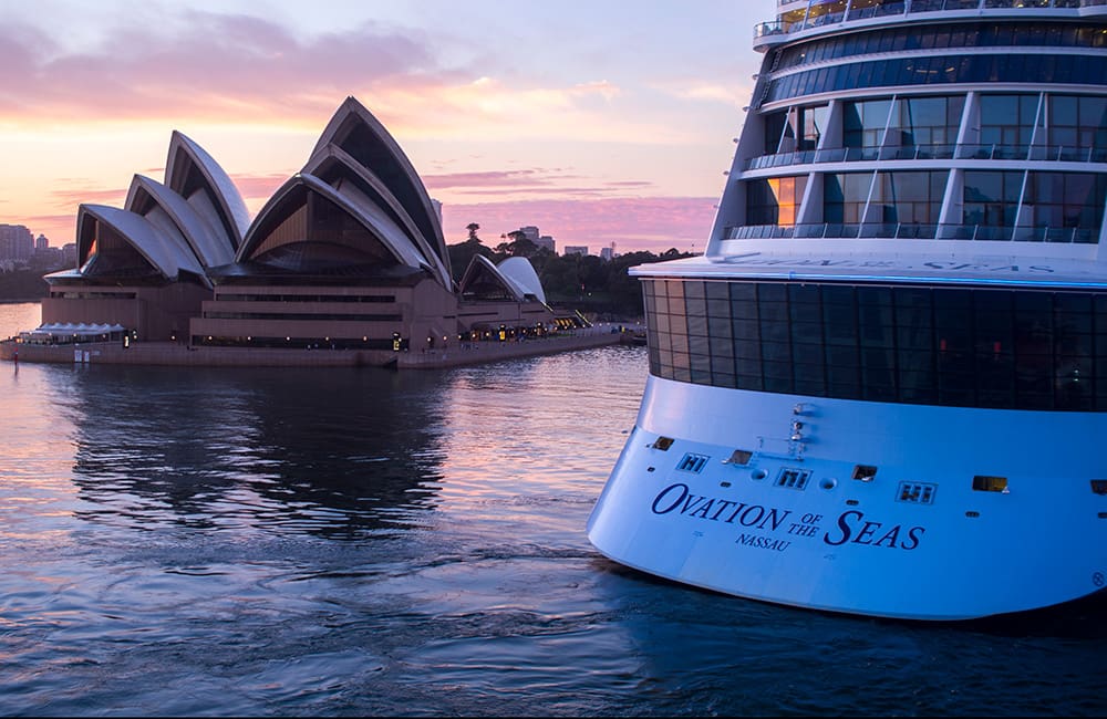 Stern profile of Ovation of the Seas in Sydney Harbour at sunset with Sydney Opera House in background.