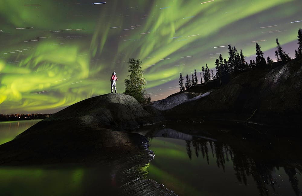 Person standing on rock at night looking at the Northern Lights in the Northwest Territories, Canada is a winter wonderland. Image: James MacKenzie