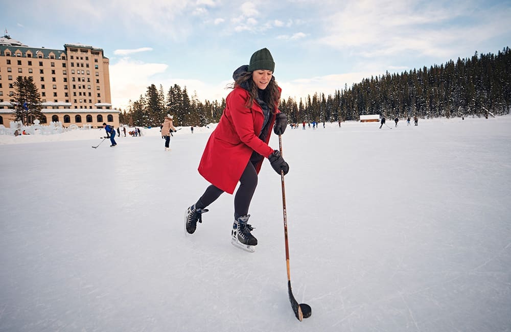 Woman and others ice skating with hockey sticks on frozen Lake Louise with Fairmont Chateau Lake Louise hotel in background. Image: Kamil Bialous