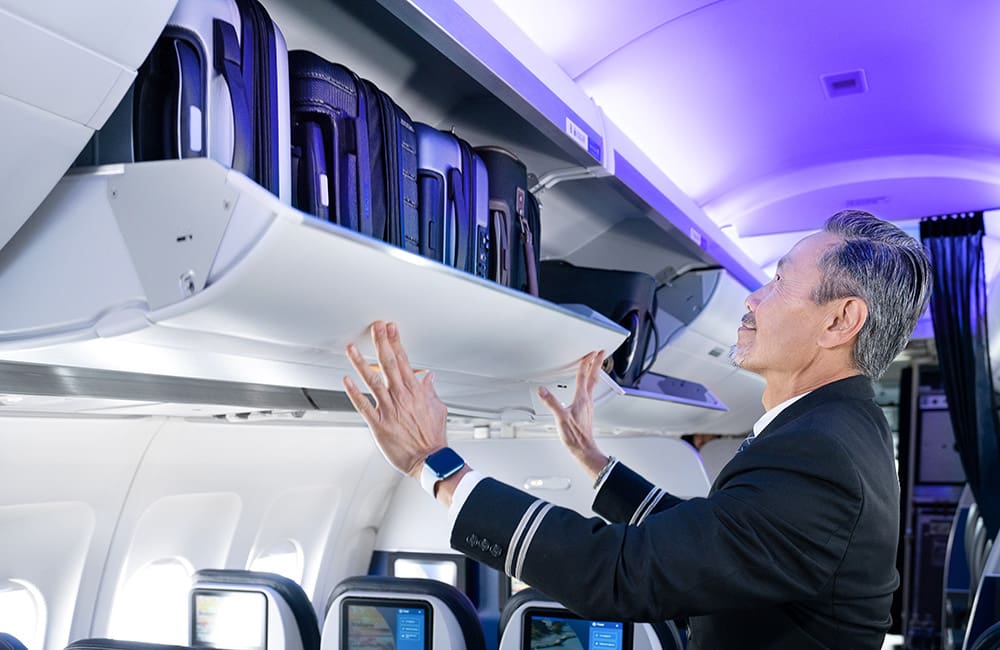 United Airlines male flight attendant closing overhead bin on plane.