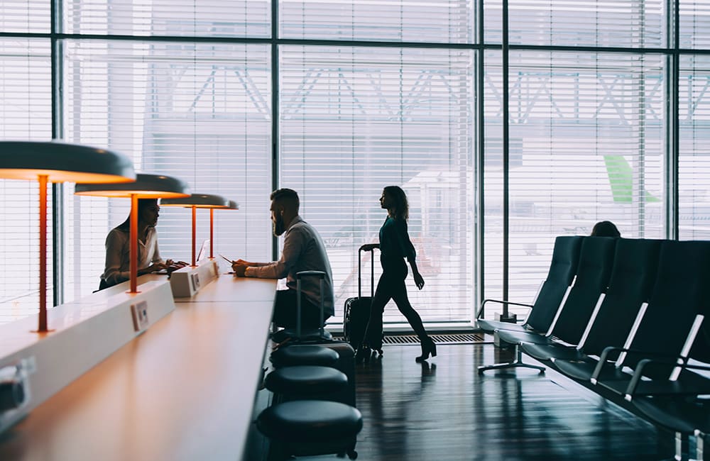 Female traveller wheeling luggage through airport with other passengers seated at desk. Image: EyeEm